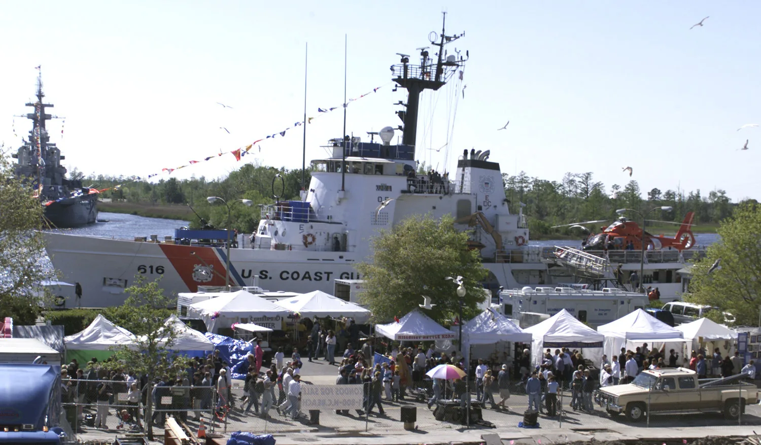 Azalea Festival crowds and booths on the Wilmington riverfront, with a Coast Guard cutter docked and the Battleship North Carolina across the Cape Fear River
